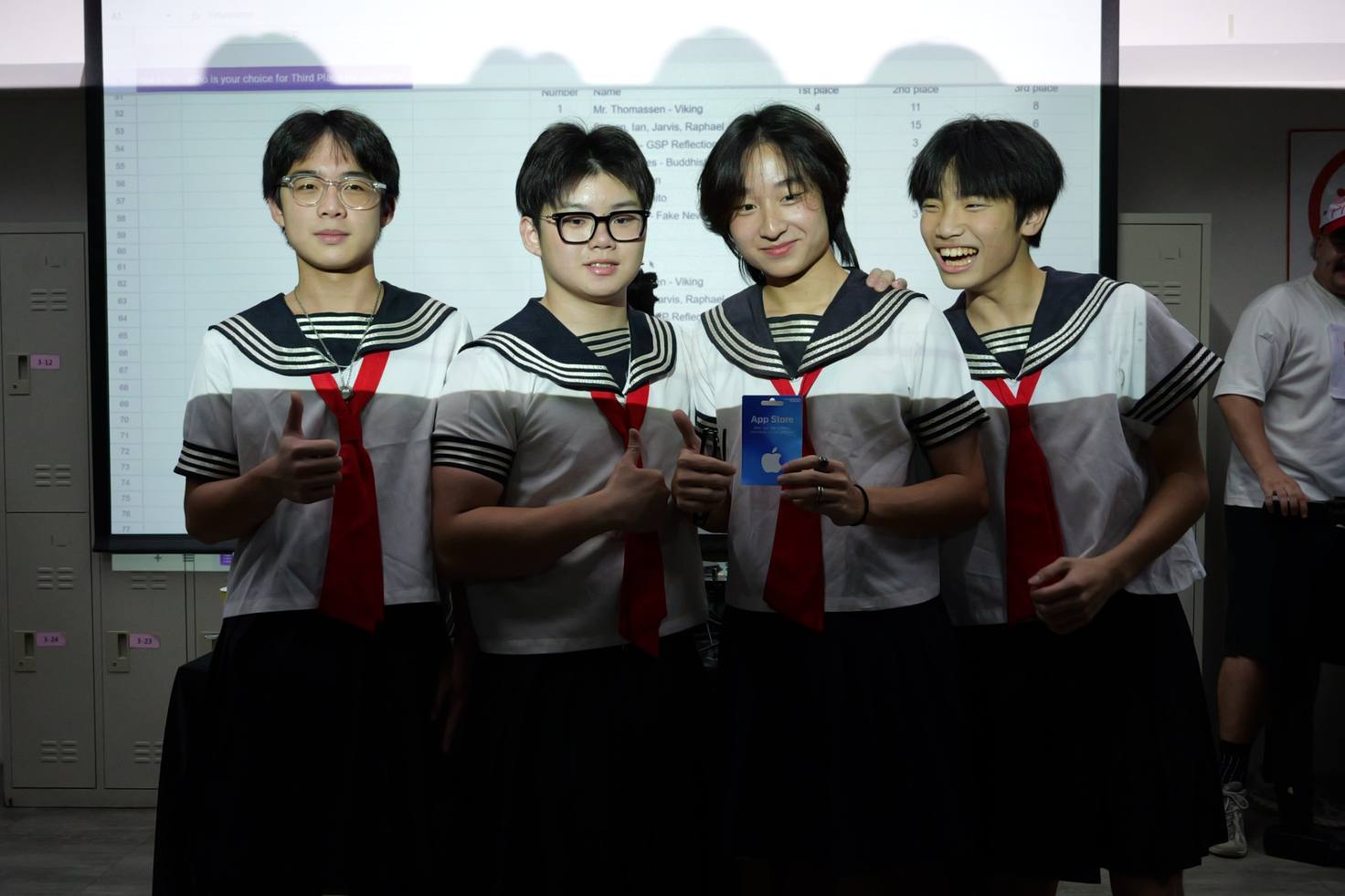 Four students in matching sailor-collar uniforms pose shoulder to shoulder; two give a thumbs up and one holds up a gift card.
