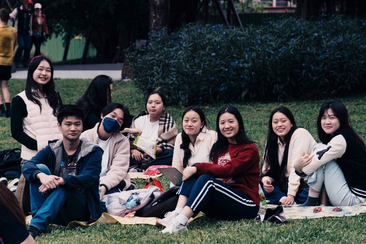 About ten students sitting together on a picnic blanket on the grass, turned towards the camera.