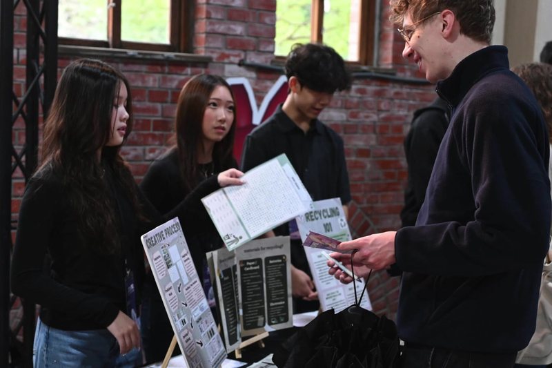 A student at a stall hands a printed leaflet to a visitor, with project boards about recycling behind them.