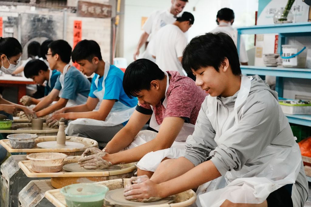 A row of students at pottery wheels, hands shaping wet clay, with a bowl of water beside each wheel.